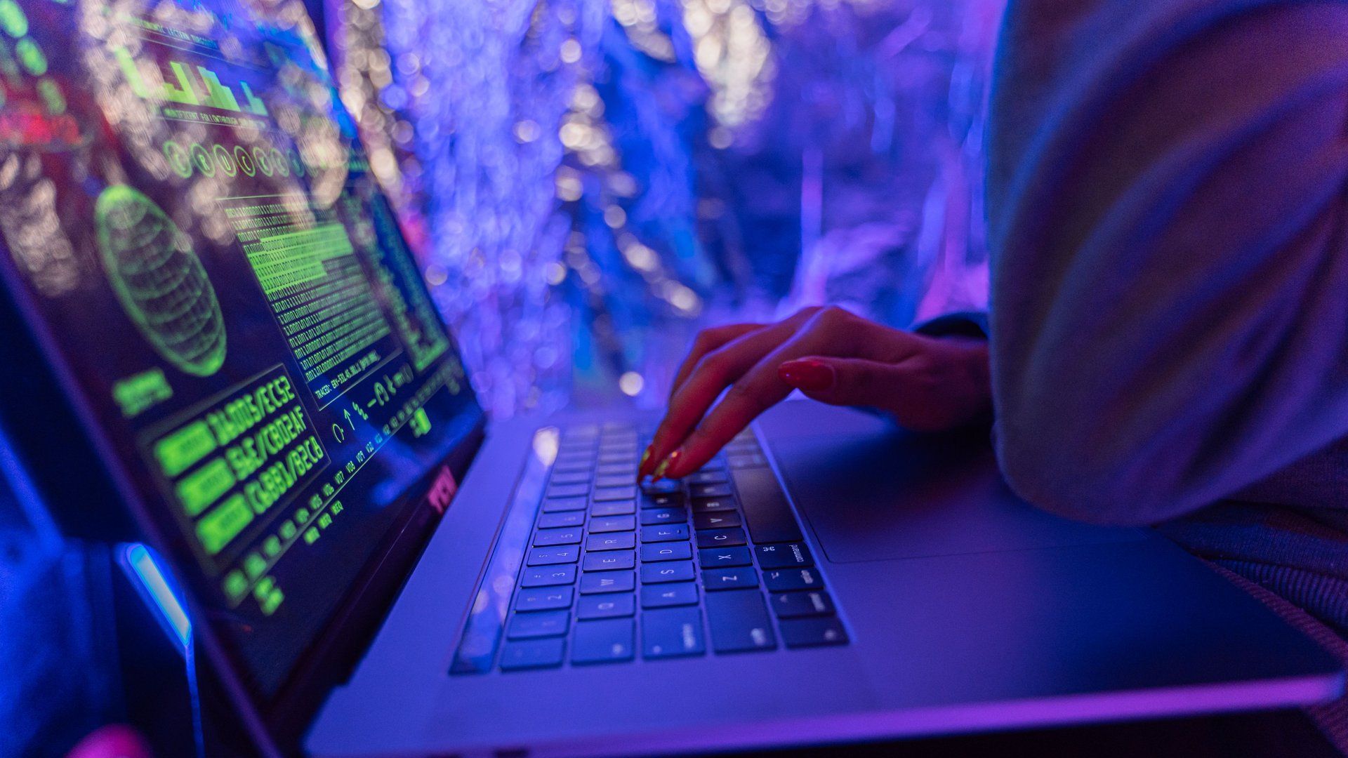 A person typing on a laptop showing a glowing data readout, in a moody blue-lit setting